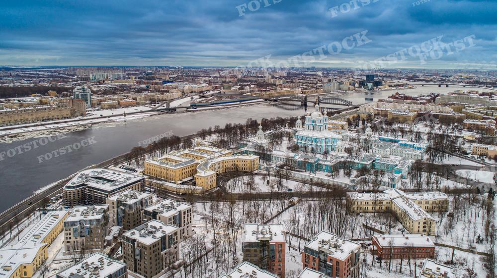 На фотографии представлен зимний вид Санкт-Петербурга с высоты птичьего полёта. Видны заснеженные улицы и крыши, а также река, пересечённая мостами. Ярко выделяется архитектурный ансамбль Воскресенского Смольного собора, окружённого более современными зданиями. Это фото может использоваться в научных и образовательных изданиях, для иллюстрации статей о зиме в городах России, в туристических гидах и как художественный элемент в публикациях, посвящённых архитектуре.